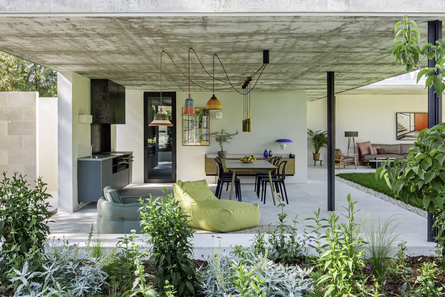 Porche de hormigón del Proyecto AS con cocina exterior, mesa de comedor, puf verde y plantas