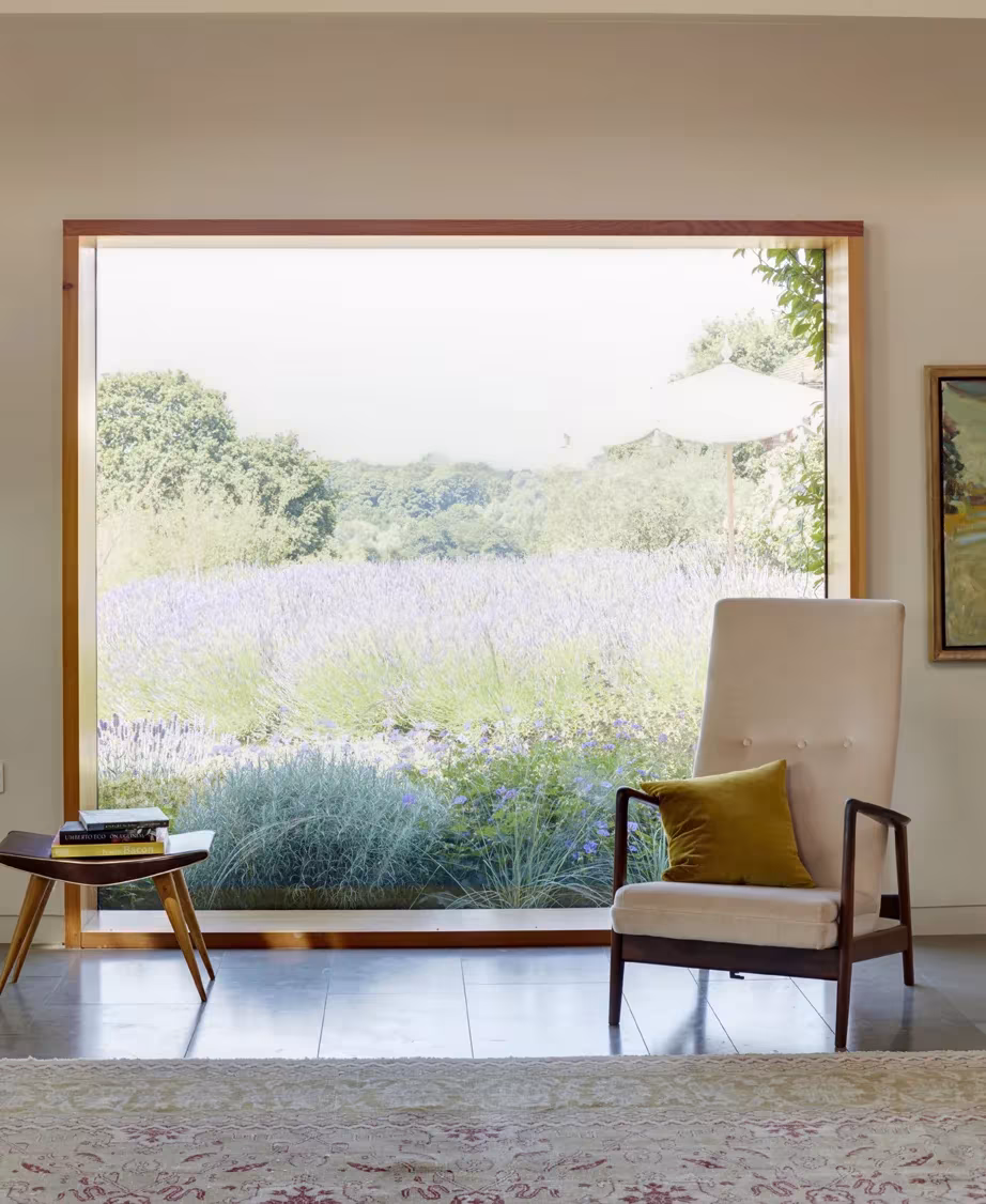 Gran ventana de Wall Farm que enmarca un campo de lavanda, con butaca tapizada y mesa auxiliar
