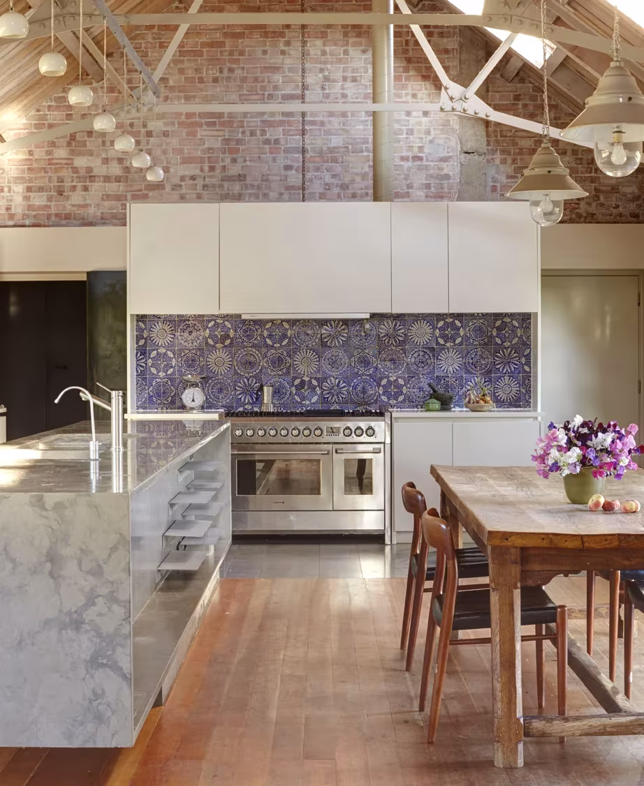 Cocina de Wall Farm bajo cerchas blancas y muro de ladrillo, con isla de mármol y mesa de madera