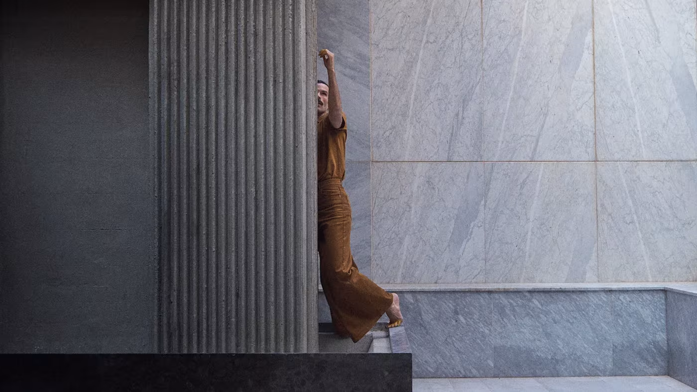 Persona con vestido ocre estirando el brazo junto a un muro de mármol gris y una columna estriada