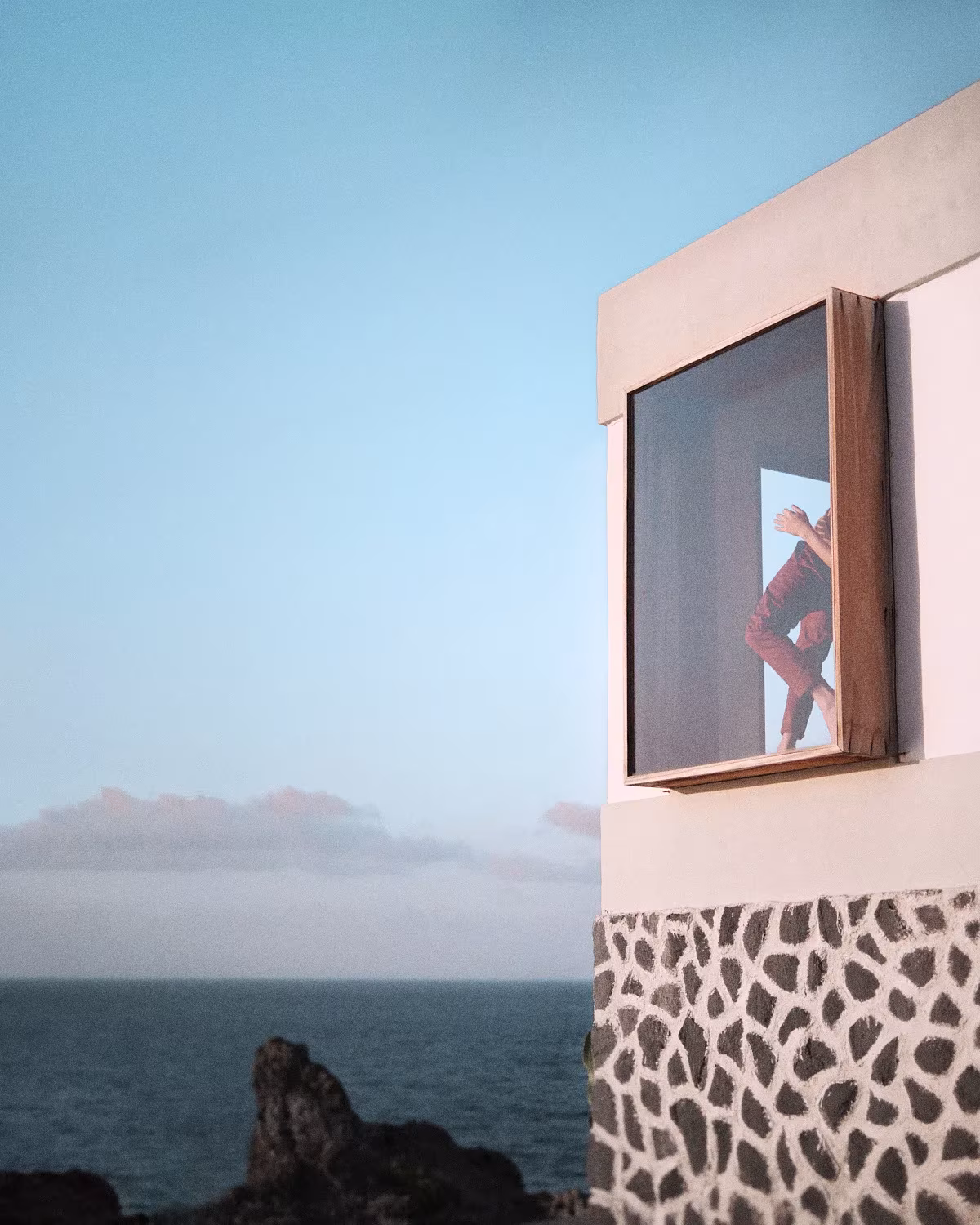 Ventana en un muro rosado con revestimiento de piedra negra sobre el mar, con una figura asomada