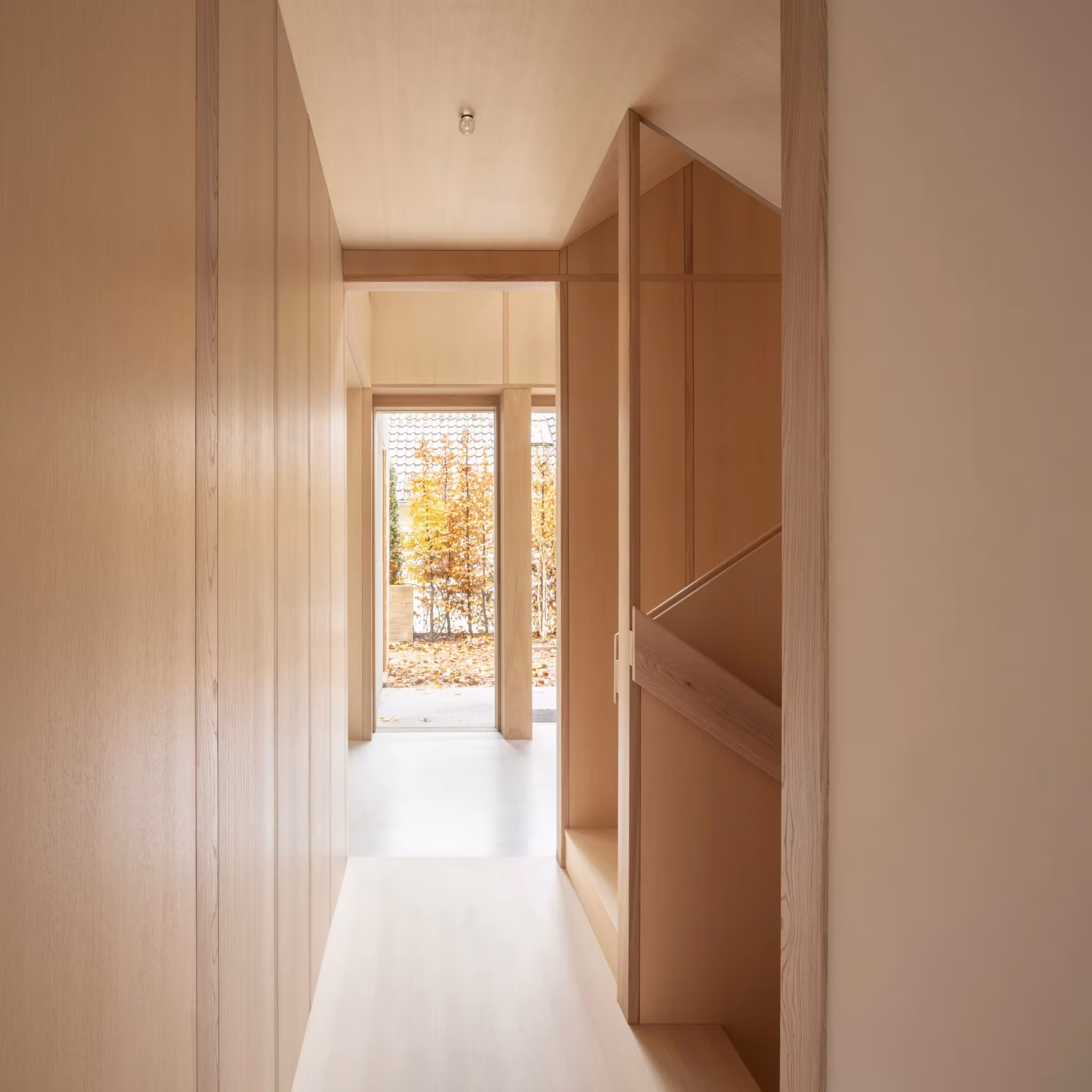 Pasillo revestido de paneles de madera clara con una ventana al fondo hacia el jardín otoñal en Fohlenweg