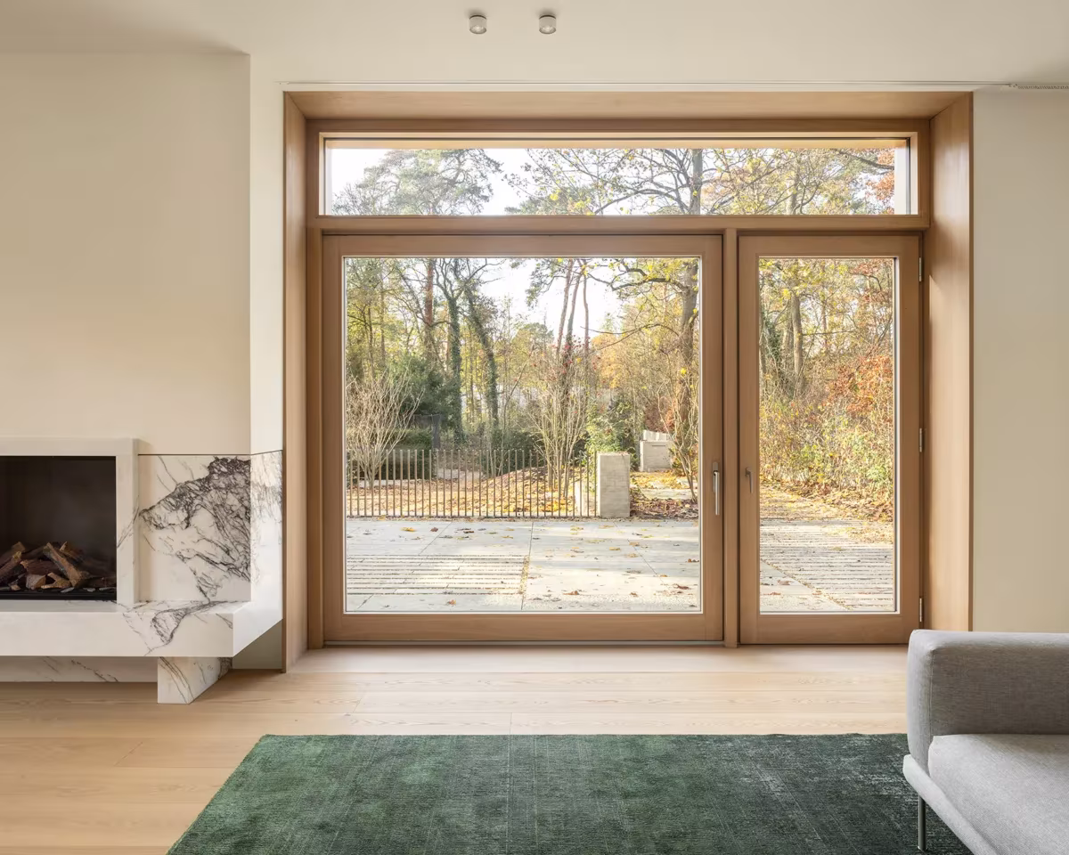 Salón de Fohlenweg con chimenea, ventanal de madera hacia el arbolado y alfombra verde