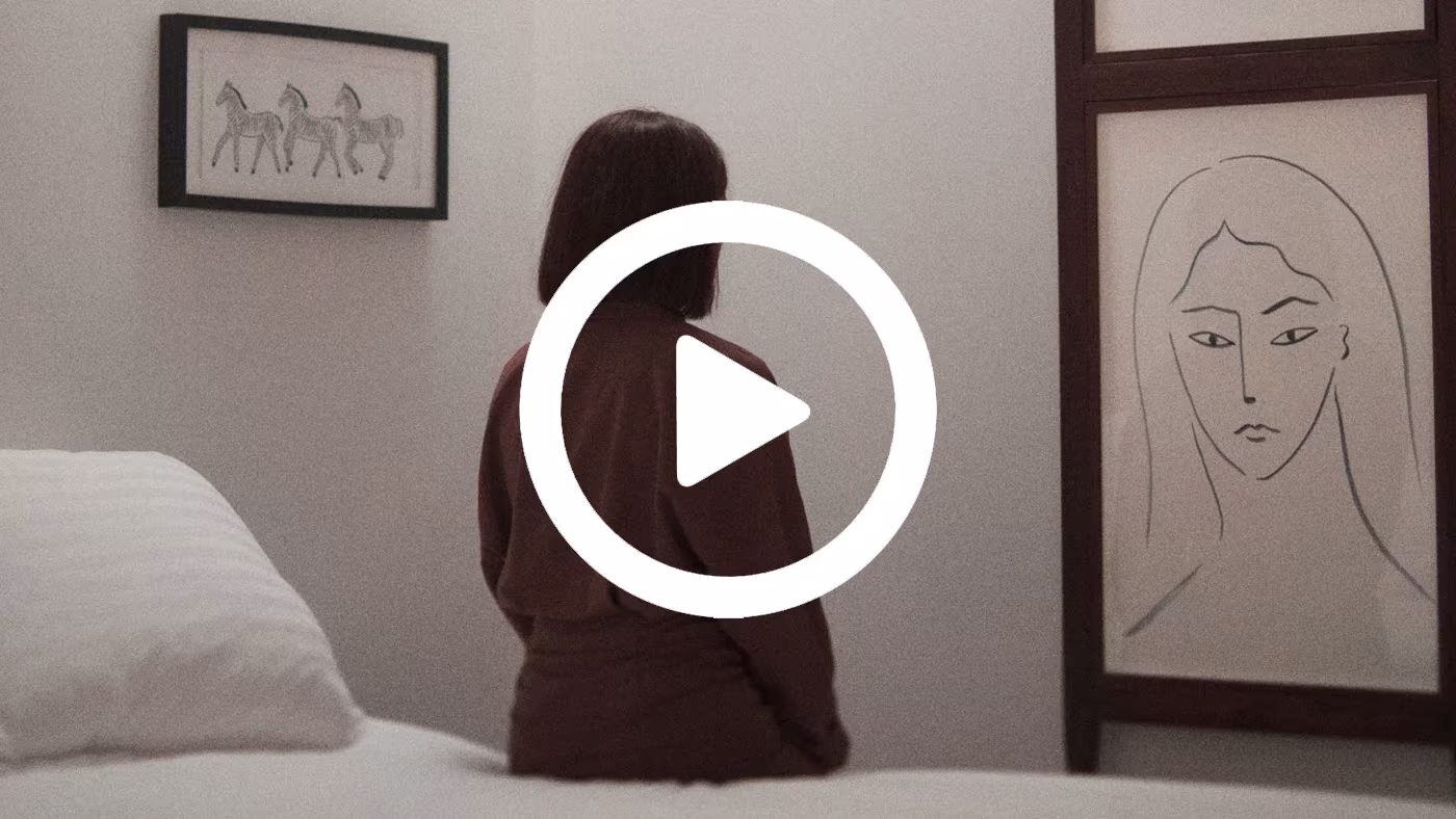Fotograma de vídeo de Derek Pedros con una mujer sentada de espaldas sobre una cama, ante un dibujo de rostro