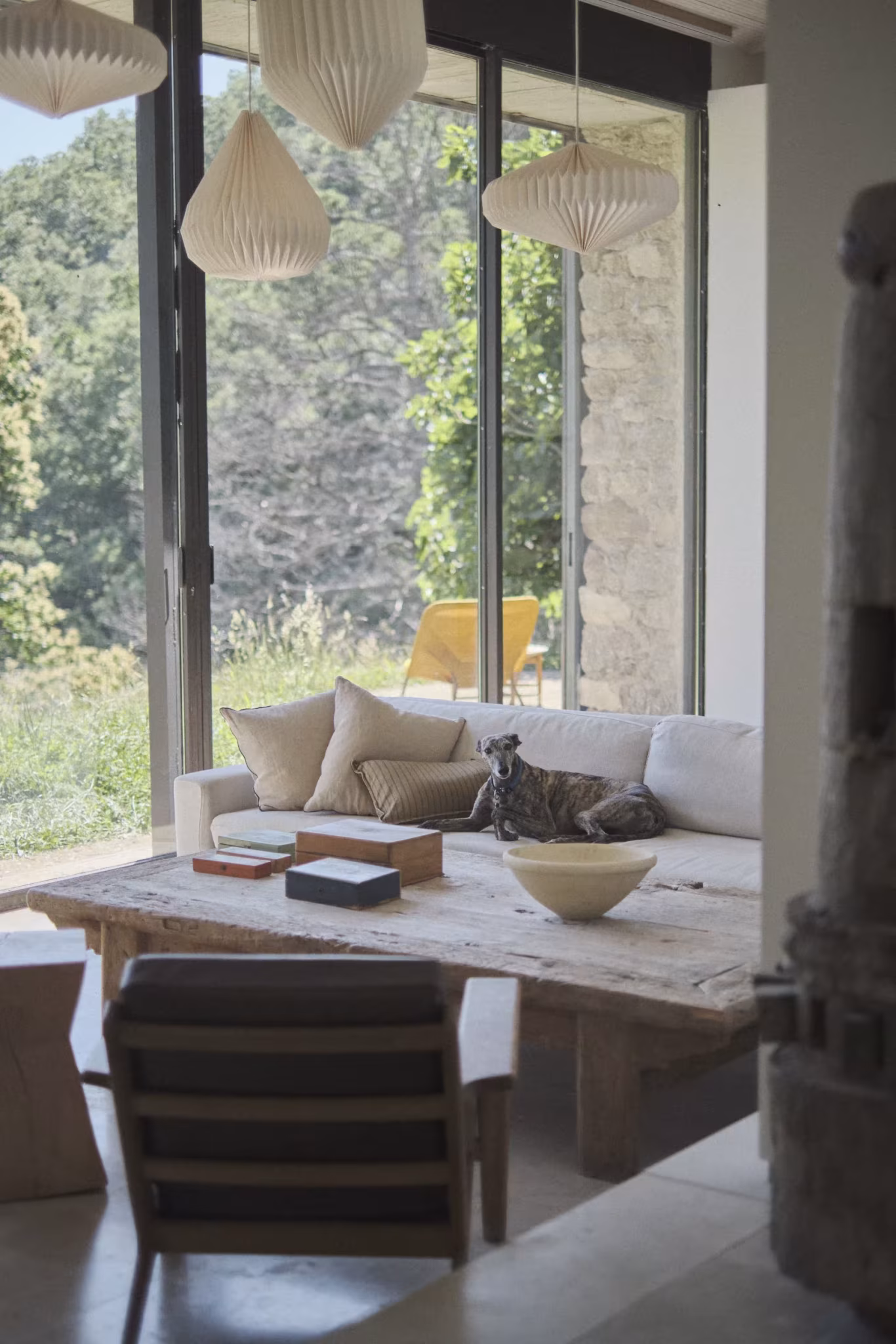 Salón con sofá, mesa baja de madera y lámparas de papel ante un gran ventanal abierto al bosque, concurso de Ábaton