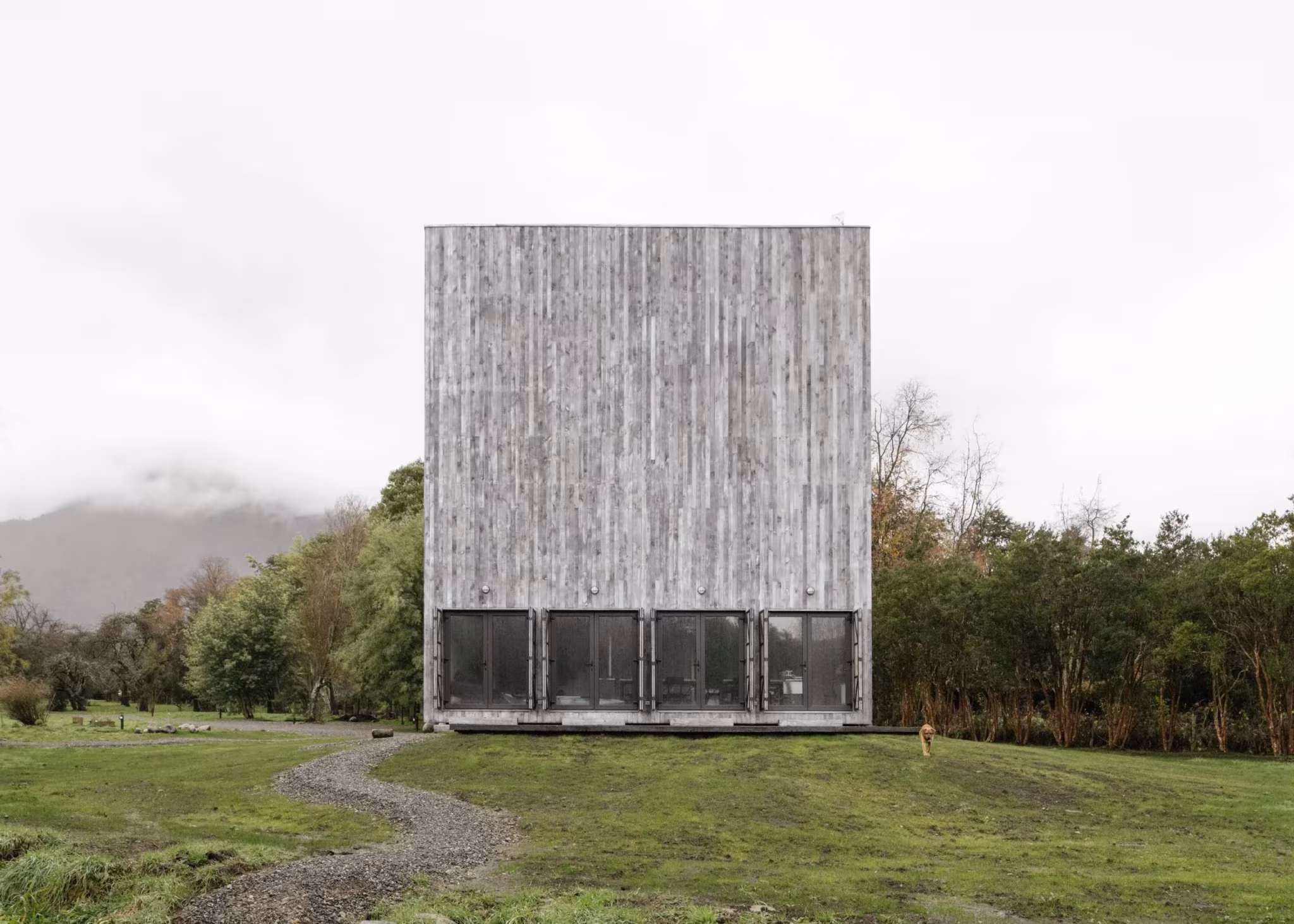 Humo House, un prisma de madera ennegrecida con base acristalada, en un prado con montañas entre la niebla