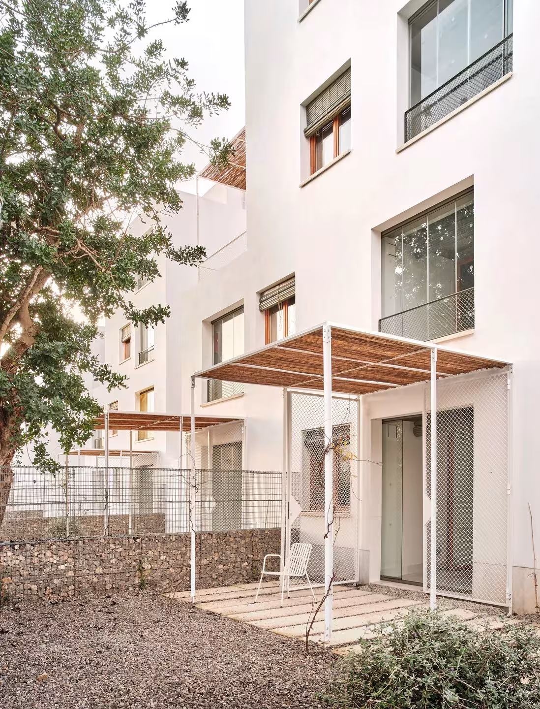 Trasera de las viviendas de Ibiza con pérgola ligera de madera sobre un patio de grava y fachada blanca