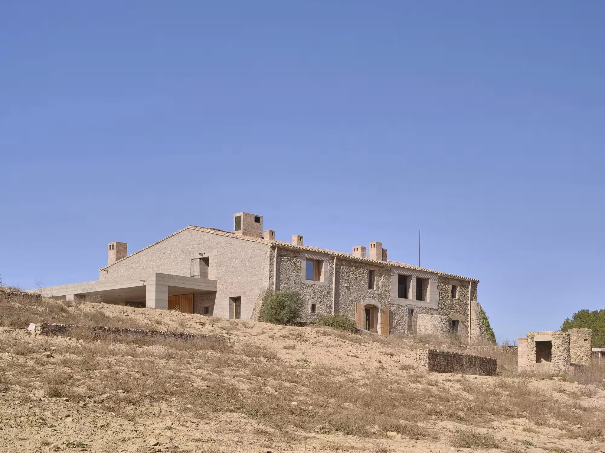 Casa 1627 de H Arquitectes vista desde la ladera, con la masía de piedra y el nuevo cuerpo de piedra clara