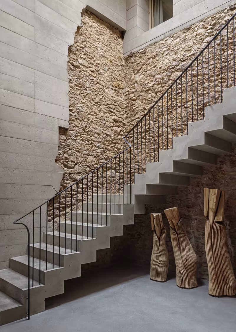 Escalera de hormigón con barandilla metálica y esculturas de madera junto al muro de piedra en la Casa 1627