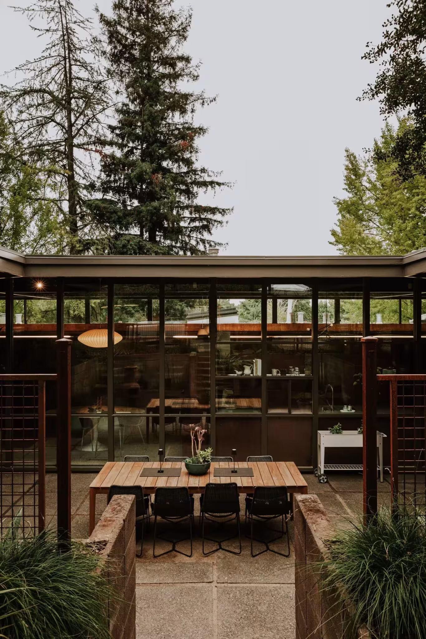 Patio con mesa de madera y fachada acristalada de la Residencia Nittler bajo los abetos, Sacramento
