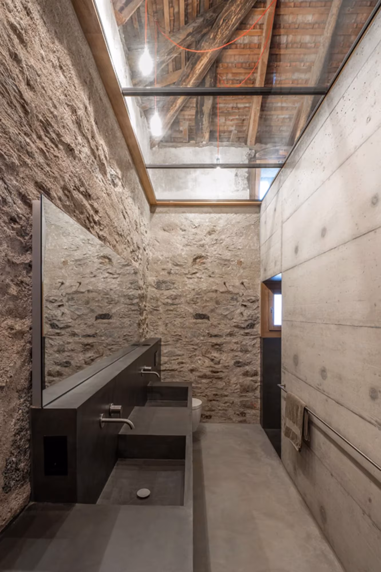 Baño con lavabo corrido de piedra oscura, muro de hormigón y lucernario en Transformation House ME