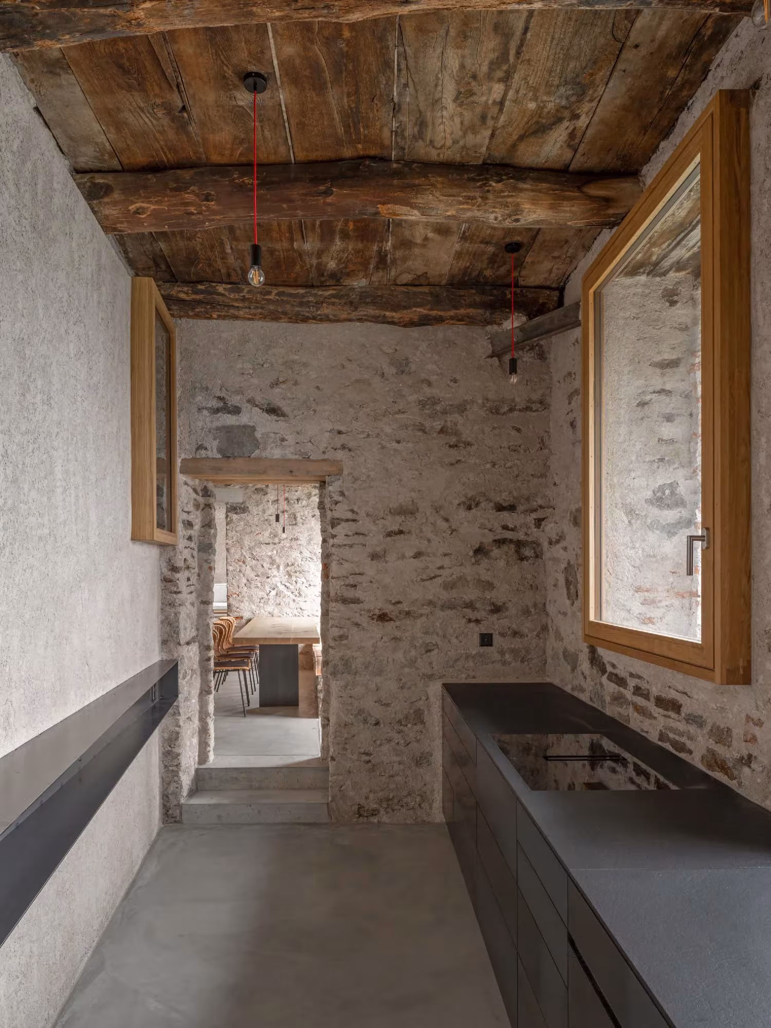 Cocina larga de encimera oscura y ventana grande entre muros de piedra y techo de madera en Transformation House ME