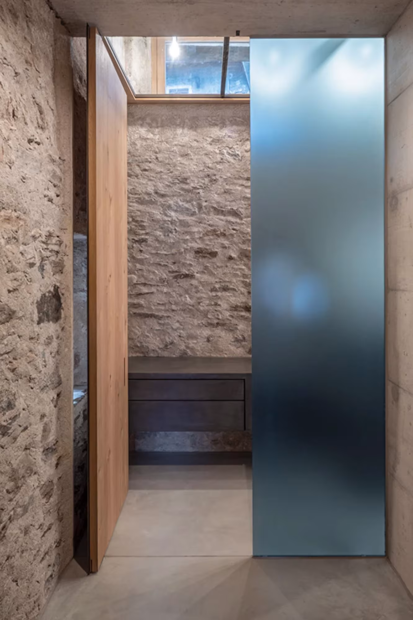Panel de vidrio translúcido azulado junto a una puerta de madera ante un muro de piedra en Transformation House ME