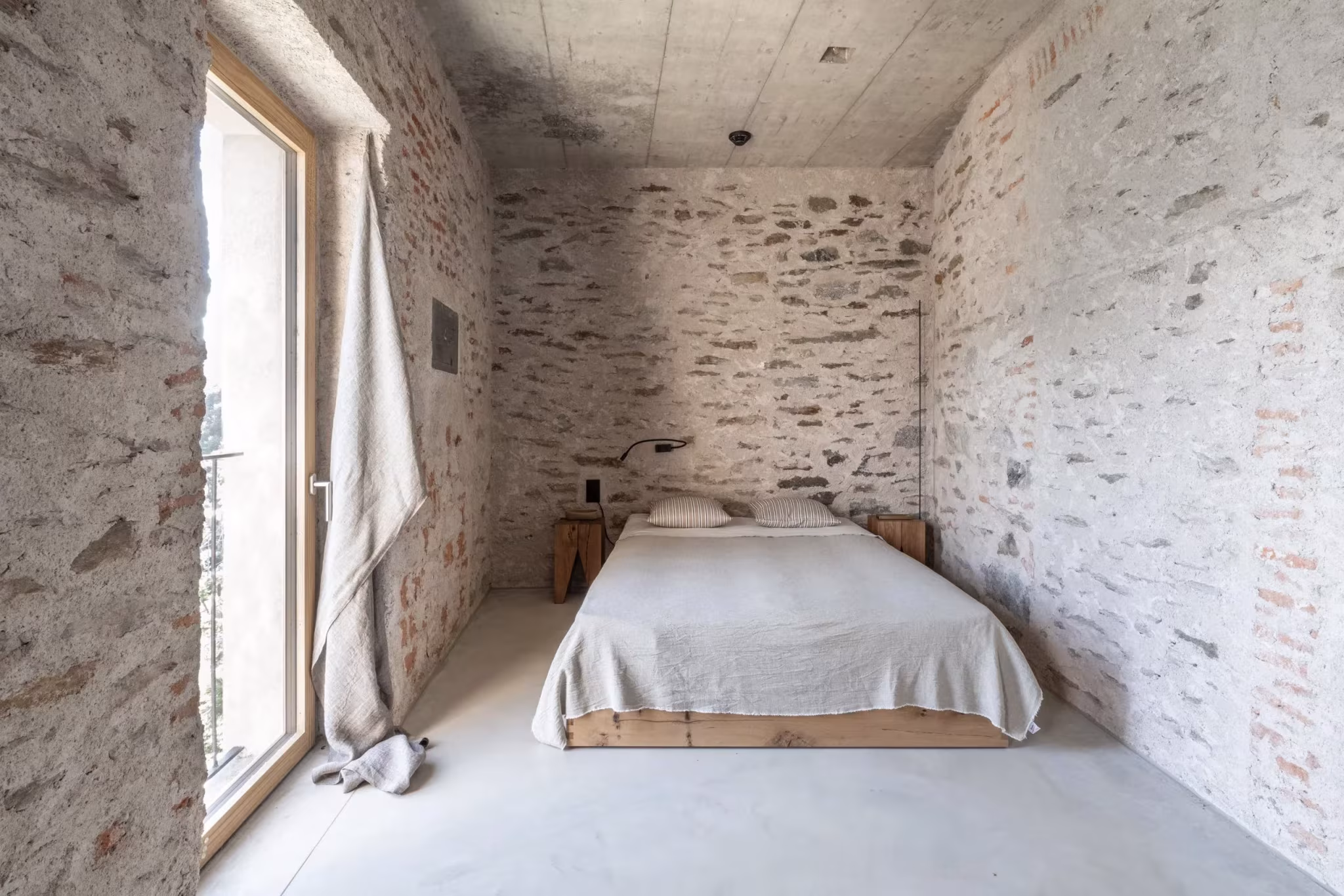 Dormitorio de suelo continuo con cama baja y ventana alta entre muros de piedra en Transformation House ME