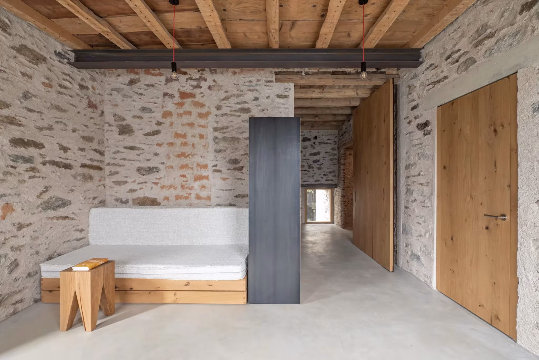 Sala con sofá bajo de madera, estructura de acero y puerta de roble ante muros de piedra en Transformation House ME