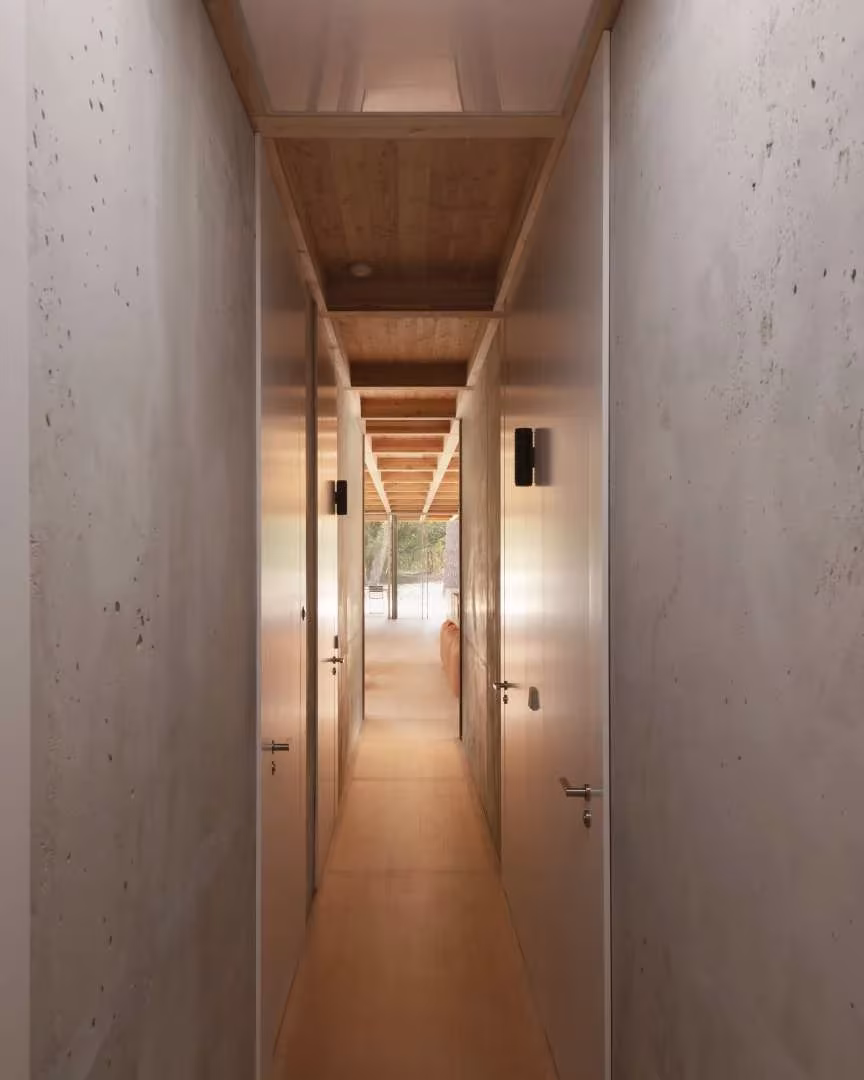 Pasillo estrecho entre muros de hormigón visto y puertas de madera con luz cenital en la Maison en Bois