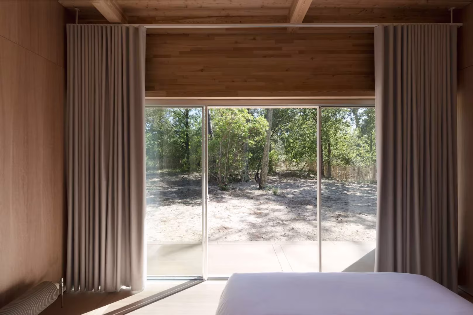 Dormitorio de la Maison en Bois con cortinas de lino ante una puerta corredera abierta al pinar