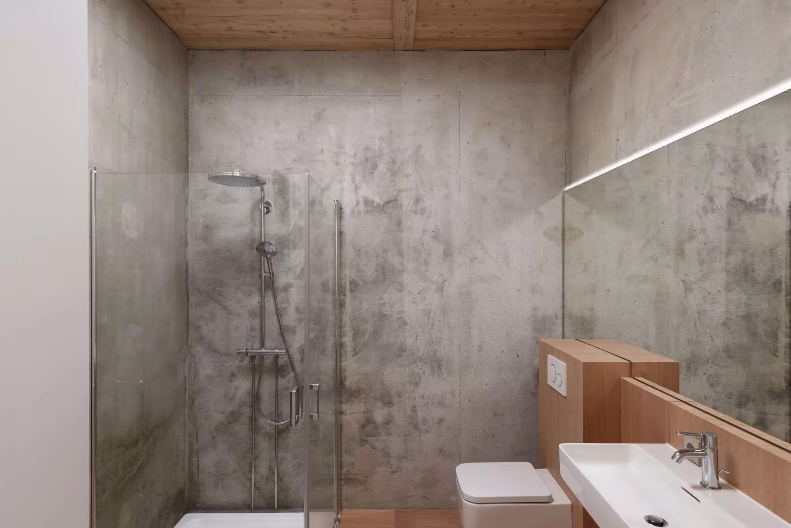 Baño de hormigón visto con ducha acristalada, lavabo y techo de madera en la Maison en Bois