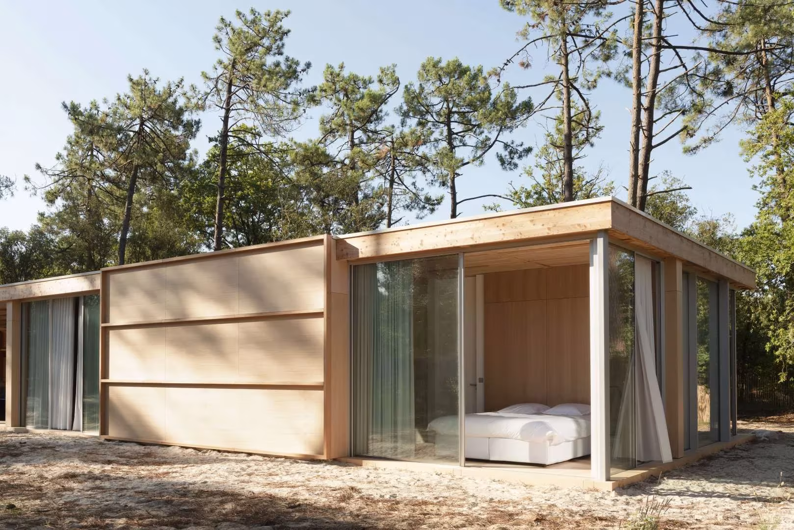 Dormitorio con cama blanca tras la cristalera en una esquina de la Maison en Bois rodeada de pinos