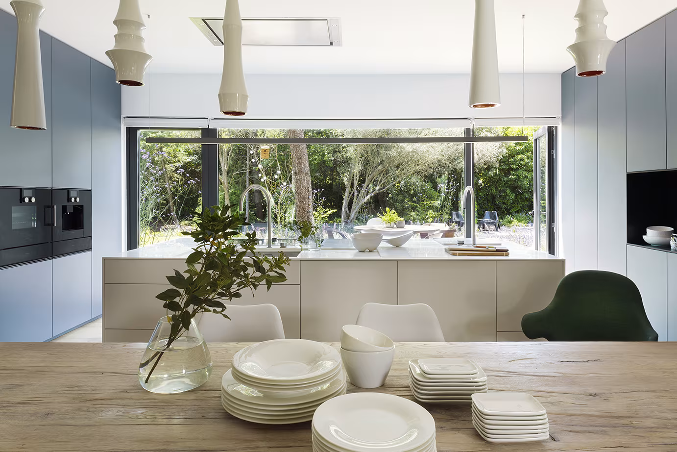 Cocina con isla y mesa de madera, lámparas colgantes y ventana corrida hacia el jardín en el Proyecto CH