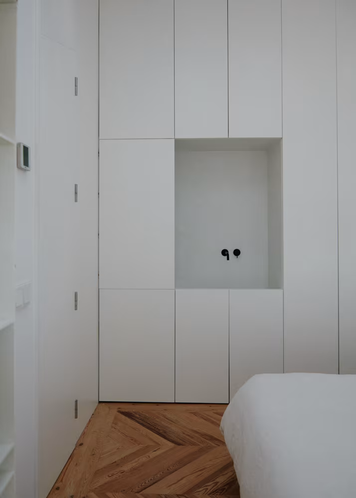 Espacio minimalista con nicho de grifería negra empotrada y cama blanca parcialmente visible