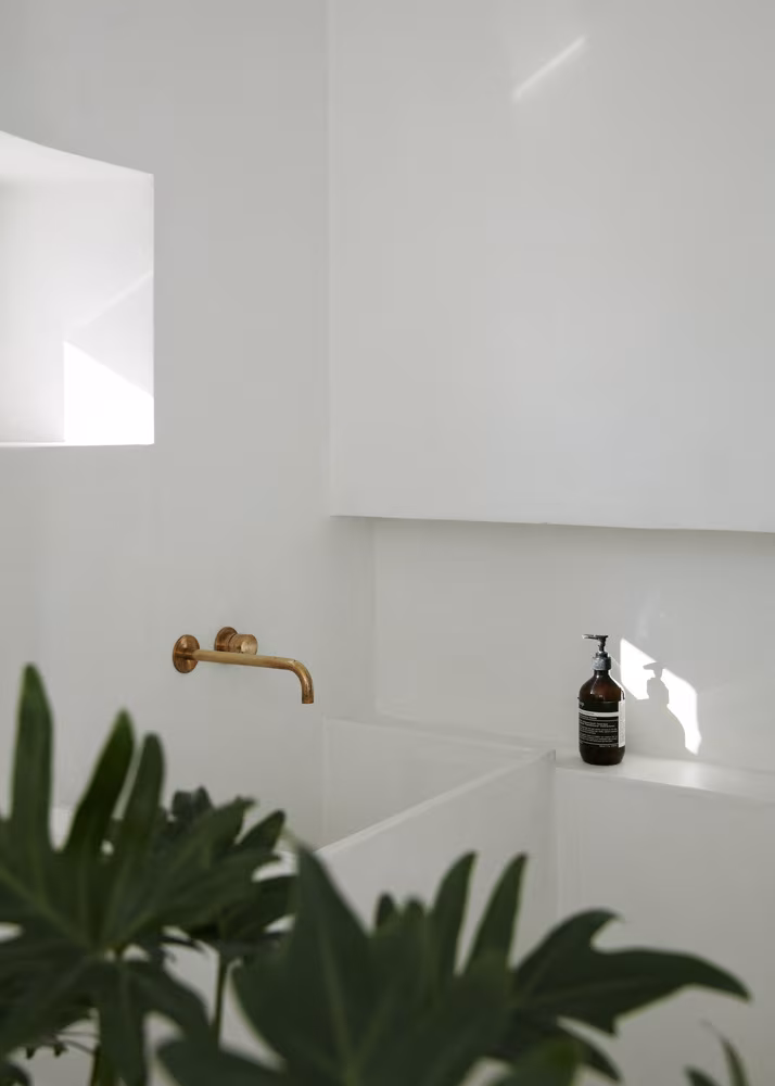 Lavabo blanco con grifería dorada, dispensador de jabón sobre encimera clara, ventana lateral que aporta luz natural y hojas verdes en primer plano.
