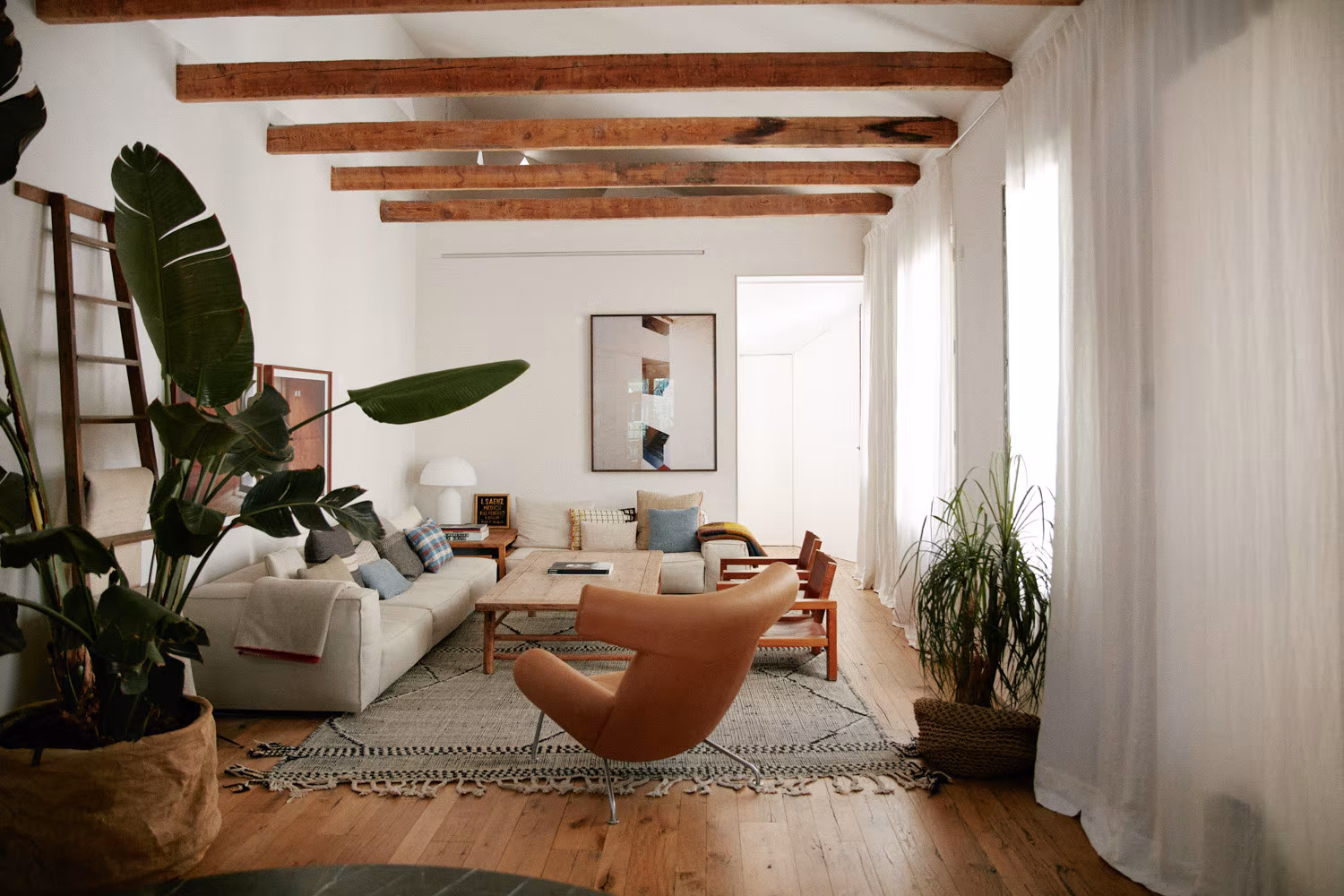 Living room with timber beams, pale sofa, leather armchair and white curtains before tall windows