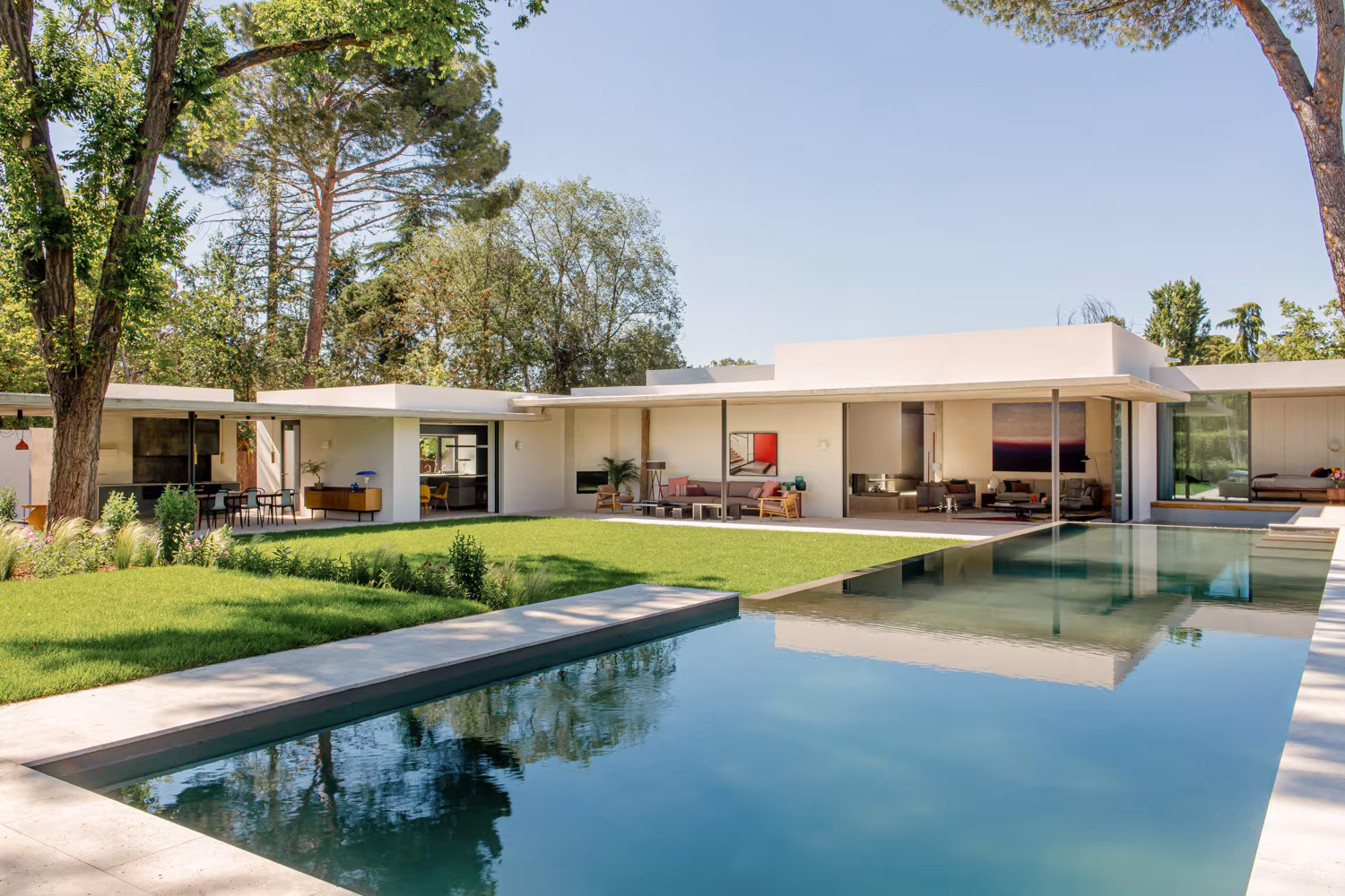 Casa moderna de una planta con fachada blanca, grandes ventanales, porche amueblado y piscina infinita rodeada de jardín, integrada en un entorno natural con árboles altos y césped bien cuidado.
