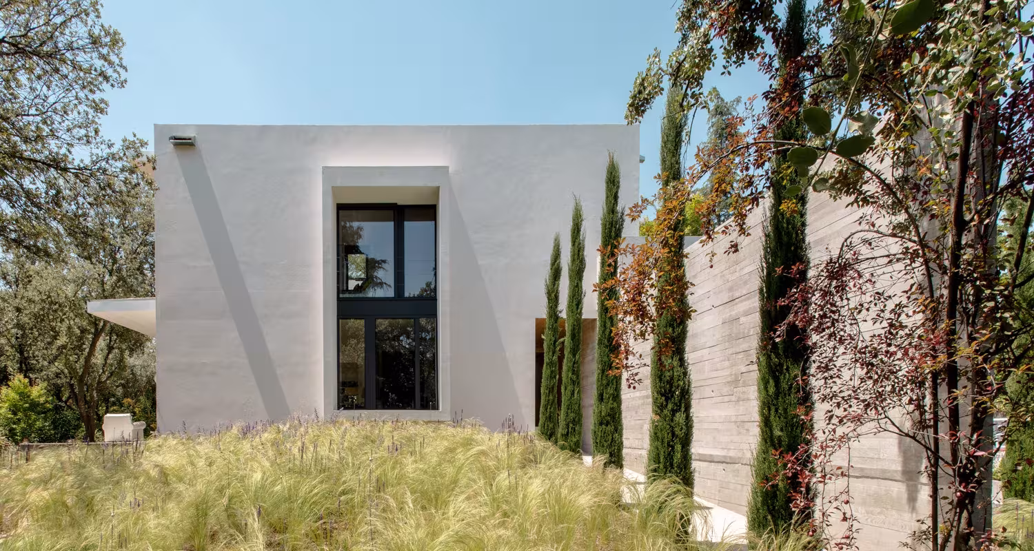 Volumen blanco con ventana vertical entre cipreses y hierbas ornamentales en el Proyecto CH de Ábaton