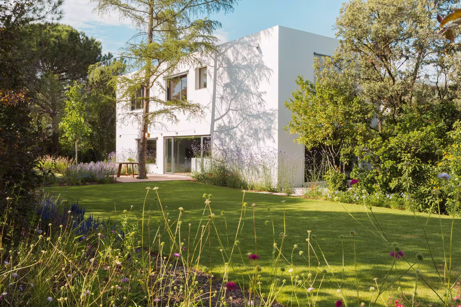 Casa blanca de dos plantas rodeada de césped y plantación silvestre en el proyecto de paisajismo CH
