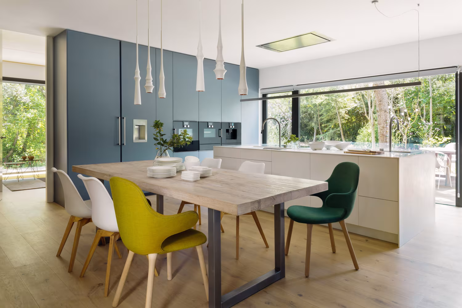 Cocina-comedor con mesa de roble, sillas de colores y armarios azul petróleo abierta al jardín en el Proyecto CH