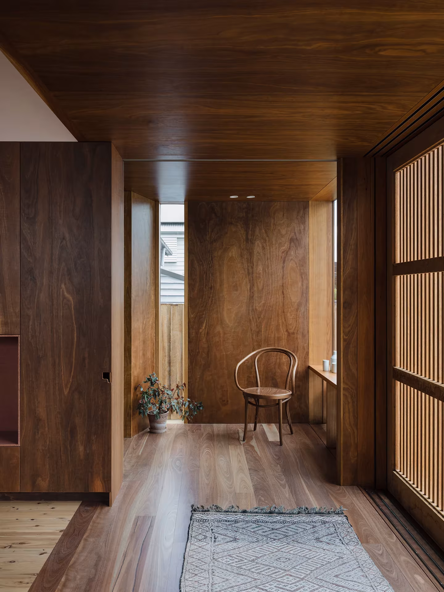 Estancia interior de madera oscura de la Aru House con ventanas de listones y un banco junto a la luz cenital