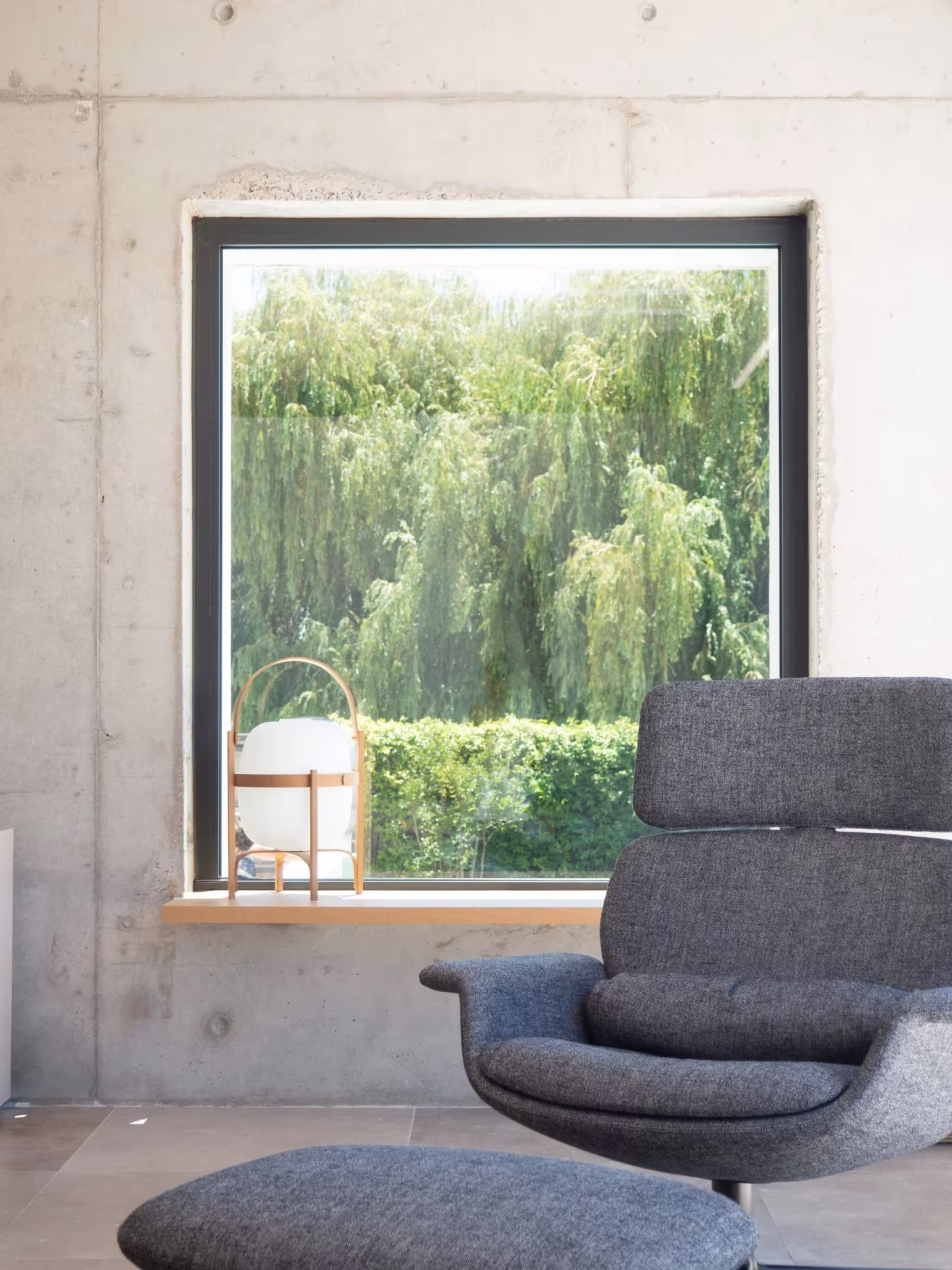 Butaca junto a una ventana cuadrada en un muro de hormigón visto con un sauce al fondo, en el artículo sobre Miguel Milá