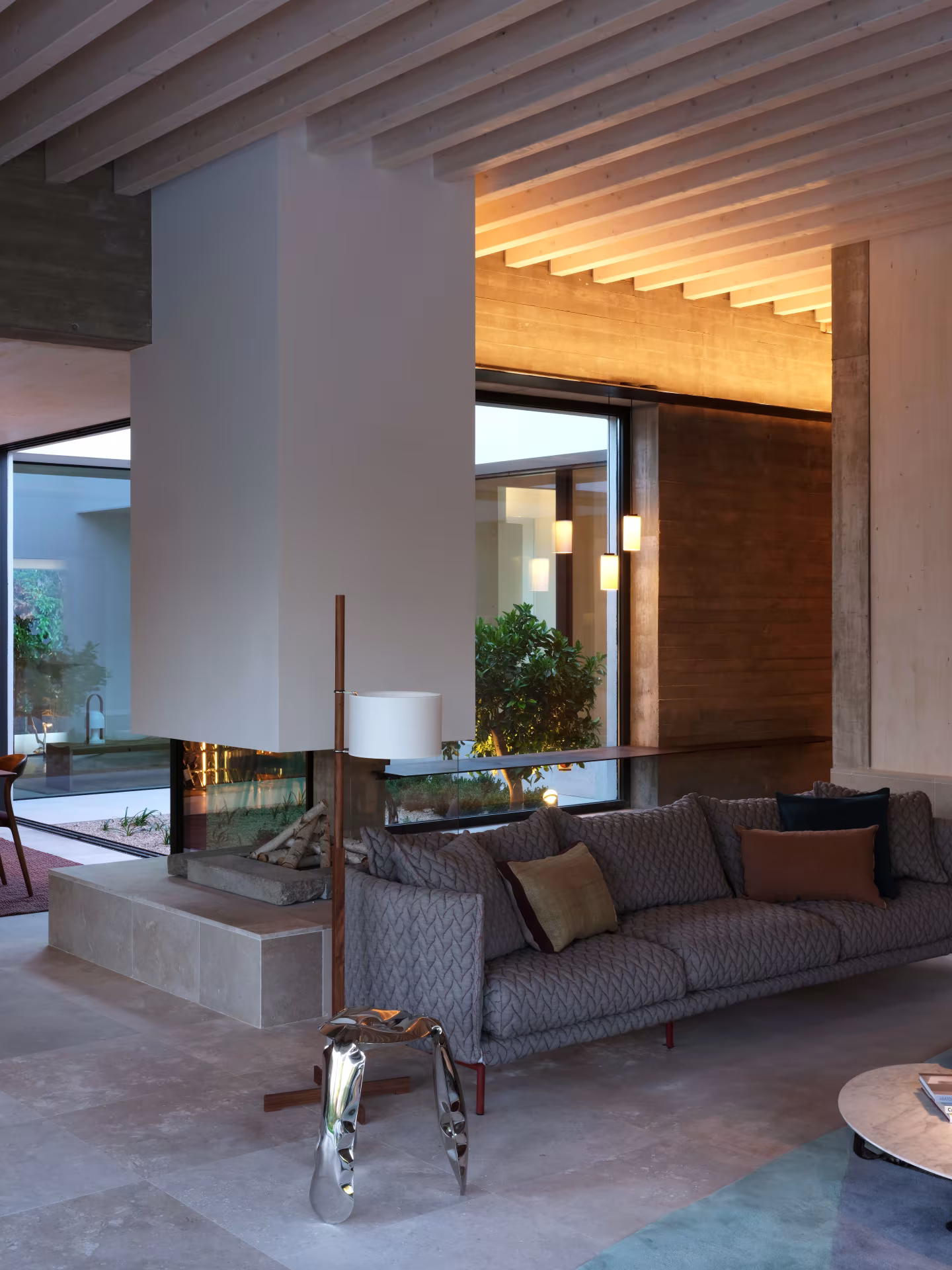 Salón con chimenea suspendida de doble cara, sofá gris y techo de vigas de madera iluminado con luz indirecta