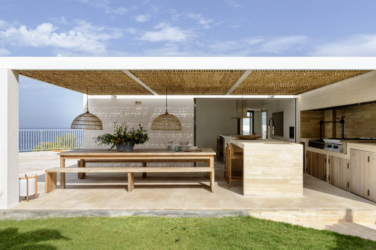 _A7B8224 Cocina exterior de estilo mediterráneo con techado de cañizo, gran mesa de comedor de madera con bancos, lámparas colgantes de fibras naturales, encimeras de piedra beige y vistas al mar bajo un cielo azul.