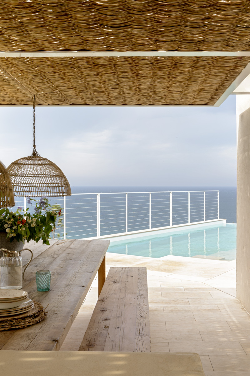 Cocina y comedor exterior resguardada bajo una pérgola artesanal de fibras naturales. Al fondo piscina infinita. 