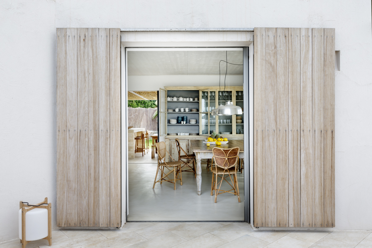 _A7B8250 Puerta corredera de madera natural abierta hacia un comedor de estilo rústico-mediterráneo, con vitrina vintage de madera envejecida, mesa de comedor robusta, sillas de fibras naturales y lámparas metálicas colgantes. Diseño interior luminoso y acogedor, conectado al exterior.