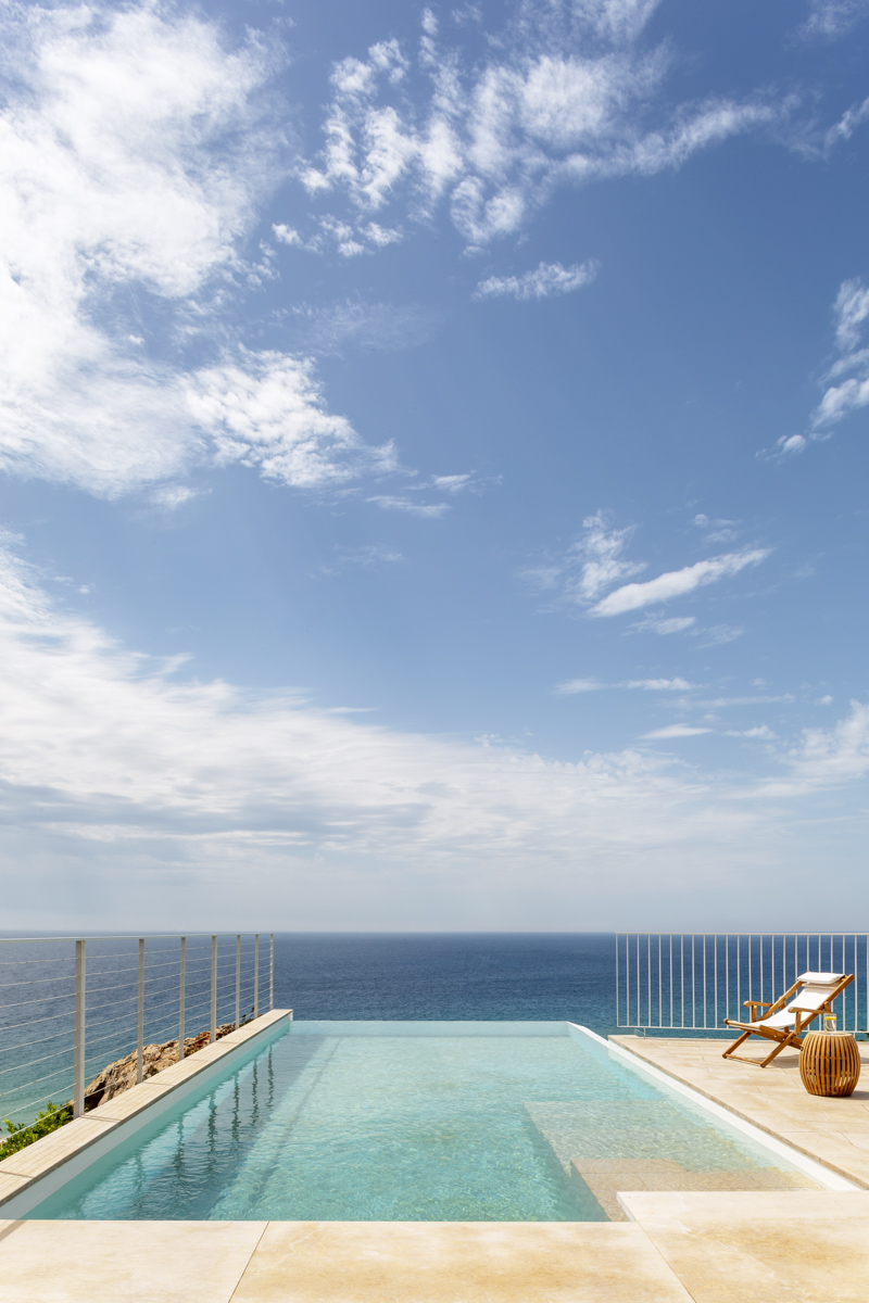_A7B8351 Piscina infinita con vistas panorámicas al mar, rodeada de barandillas minimalistas de acero inoxidable, tumbona de madera con cojín blanco y cielo parcialmente despejado en un entorno costero de lujo.