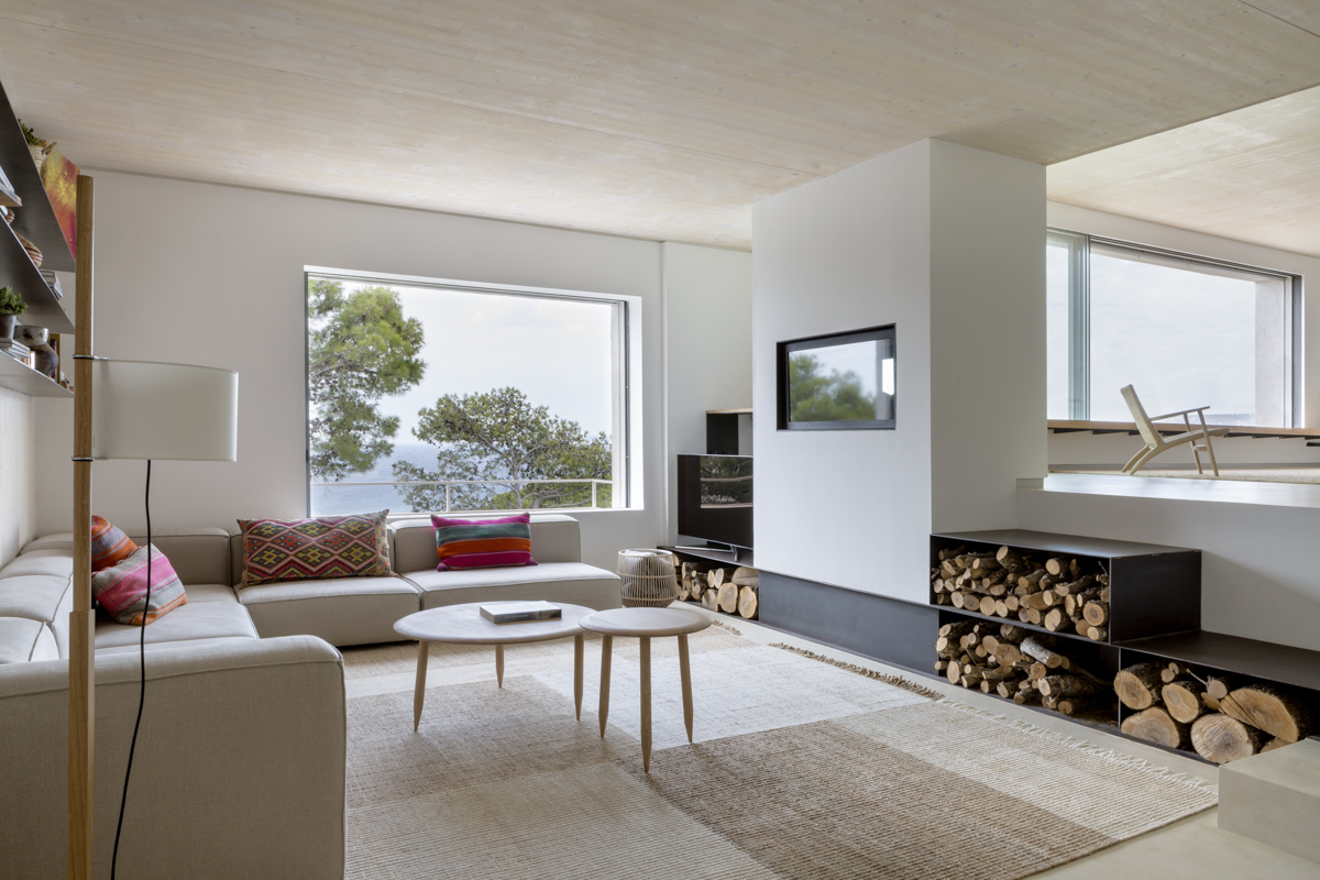 _A7B8409 Salón contemporáneo con sofá en esquina color beige, cojines con estampados étnicos, alfombra de fibras naturales, ventanales con vista al agua y naturaleza, chimenea moderna empotrada con almacenamiento de leña, y decoración minimalista con luz natural abundante.