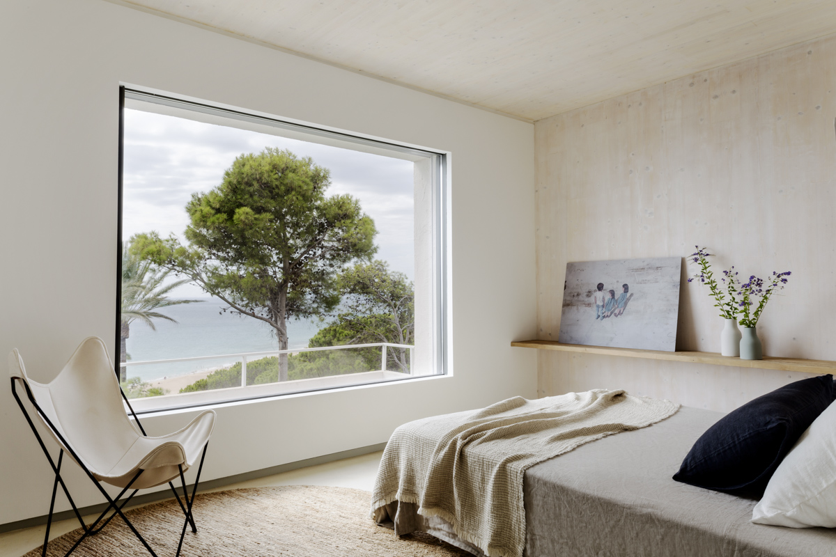_A7B8424 Dormitorio minimalista con gran ventanal panorámico con vistas al mar y árboles mediterráneos, cama con ropa de lino en tonos neutros, silla de mariposa blanca y decoración sencilla con flores frescas y arte apoyado sobre una repisa de madera natural.