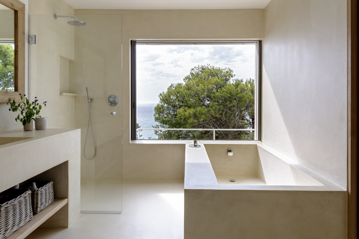 _A7B8443 Baño minimalista con ducha de cristal, bañera integrada de microcemento y gran ventana con vistas al mar y árboles. Lavabo con cestas de mimbre decorativas en el bajo