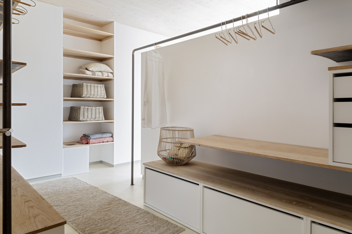 _A7B8453 Vestidor moderno de estilo minimalista con mobiliario en blanco y madera clara, estanterías abiertas con cestas de mimbre, barra de colgar ropa con perchas de madera, iluminación natural y alfombra de fibras naturales sobre suelo continuo beige.