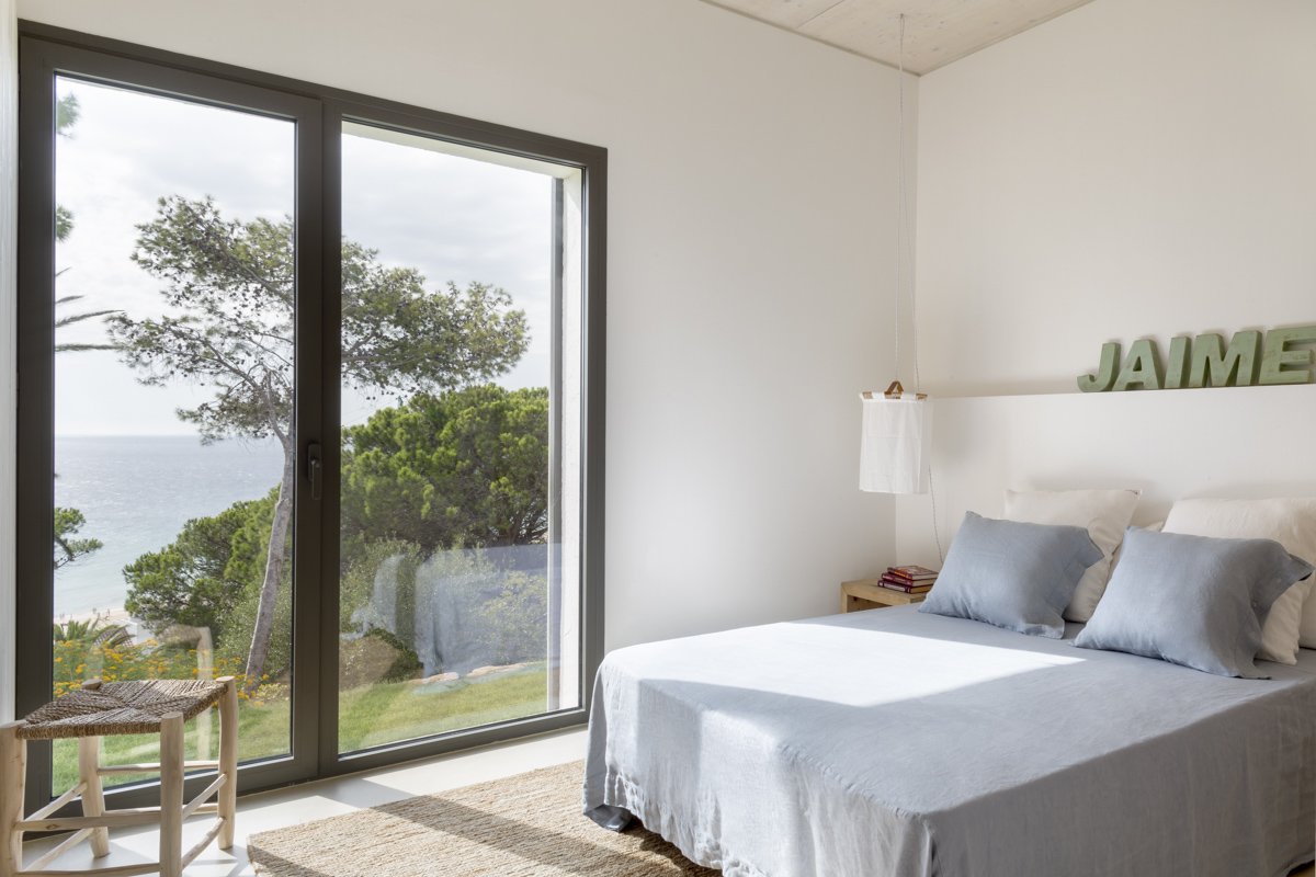 _A7B8459 Dormitorio mediterráneo con decoración minimalista y vistas al mar. Cama con ropa de cama azul claro y cojines a juego, lámpara colgante de tela, alfombra de fibras naturales y puerta de cristal con marco oscuro que da al jardín y al océano.