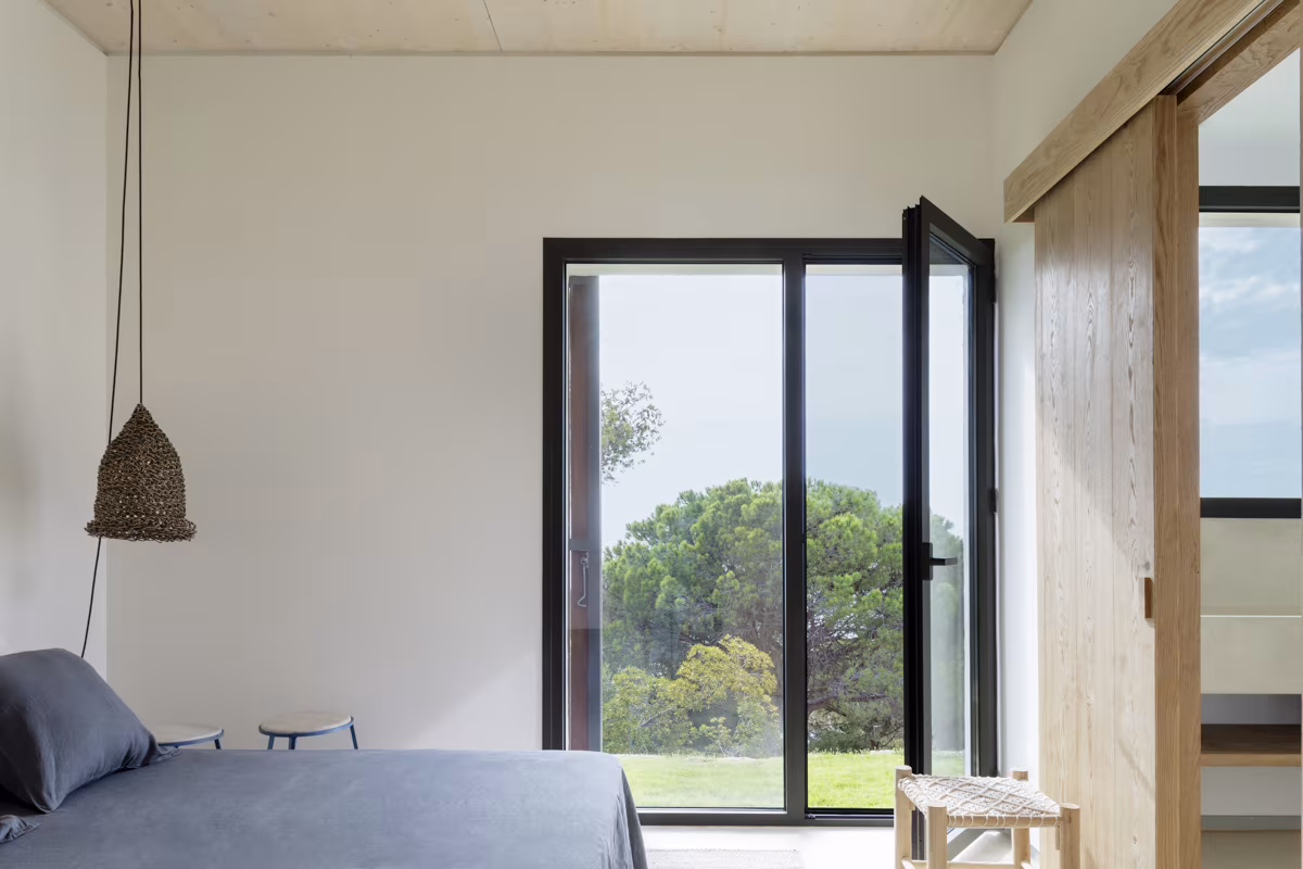 Dormitorio minimalista con ropa de cama gris y puertas de cristal de marco negro hacia el jardín
