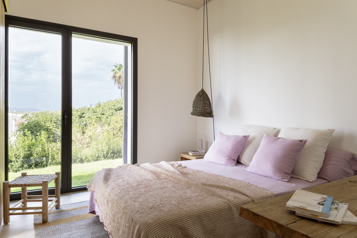 _A7B8469 Dormitorio luminoso con cama de sábanas lavanda y manta de punto, cojines blancos y rosados, lámpara colgante artesanal, y puerta con marco negro que da acceso a un jardín exterior con vegetación mediterránea.