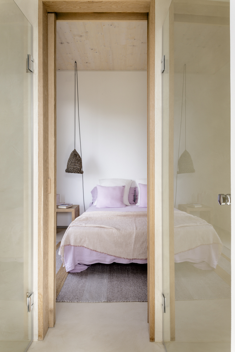_A7B8477 Vista frontal de dormitorio minimalista con cama vestida en tonos lavanda y beige, cojines suaves, lámparas colgantes artesanales y marcos de madera natural. Estilo mediterráneo con diseño acogedor y luz natural.