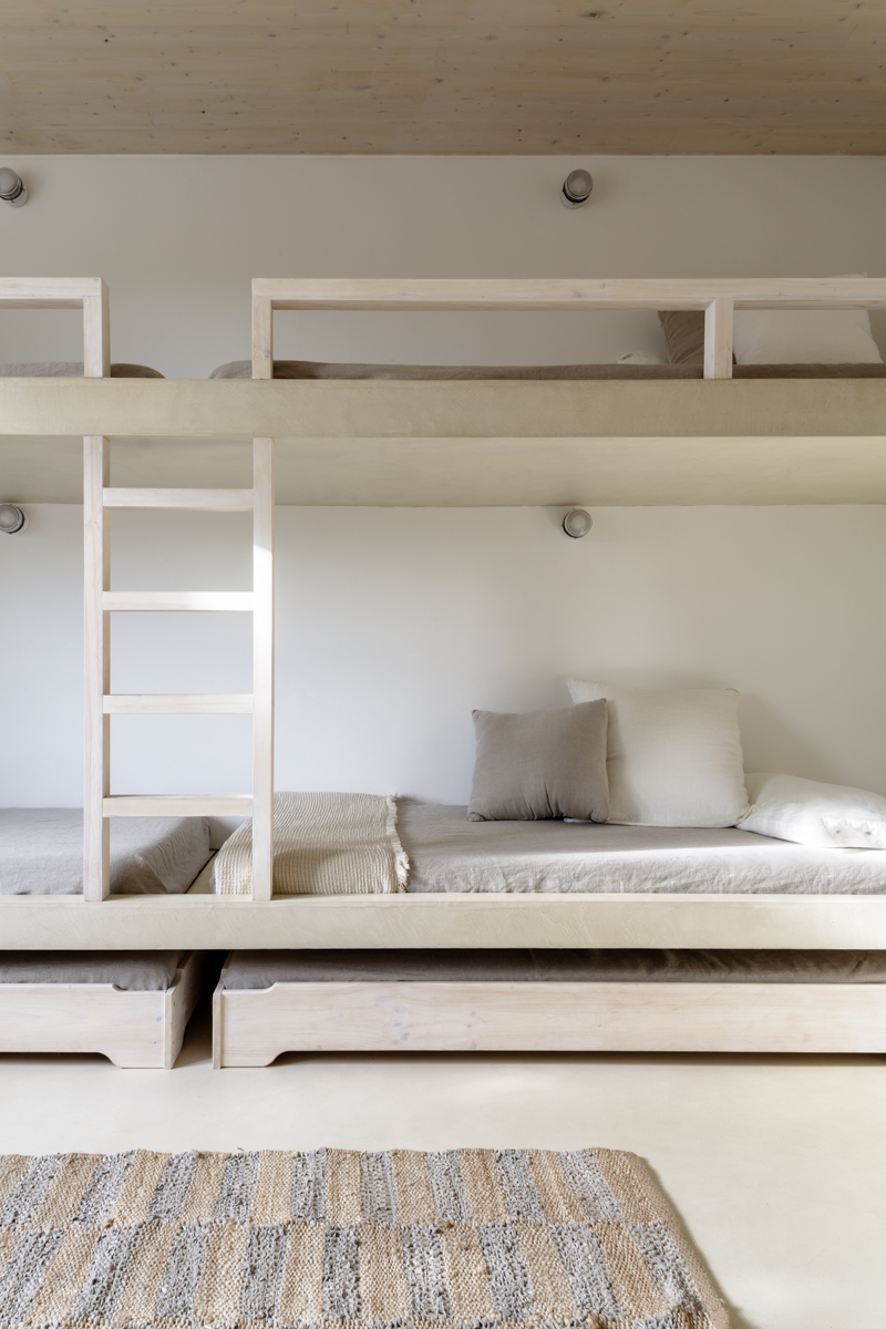 _A7B8514 Dormitorio minimalista con literas de madera clara integradas en la pared, ropa de cama en tonos beige y cojines neutros. Debajo, dos camas nido y alfombra de fibras naturales sobre suelo continuo de microcemento. Estilo mediterráneo y luminoso