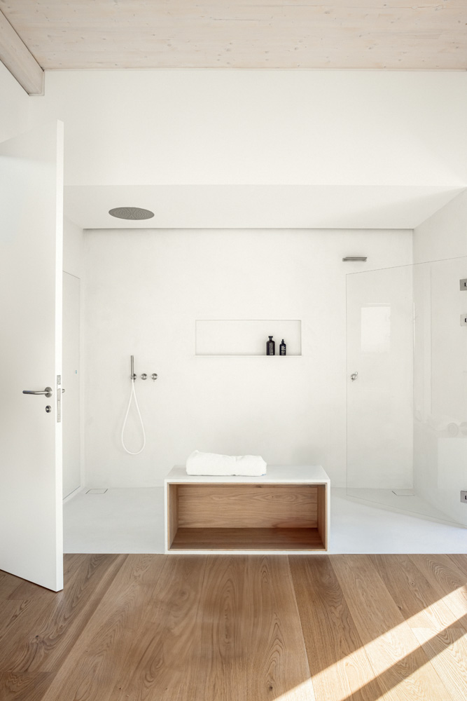 ABATON-CasaBrisa-1 Baño minimalista con ducha a ras de suelo, paredes blancas, banco de diseño con acabado en madera clara, grifería empotrada y suelo de madera natural que aporta calidez al espacio contemporáneo.