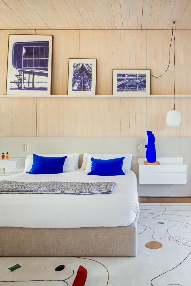 Dormitorio de diseño con cabecero de madera clara, cama doble con cojines azules vibrantes, alfombra con motivos abstractos y láminas artísticas en la pared.