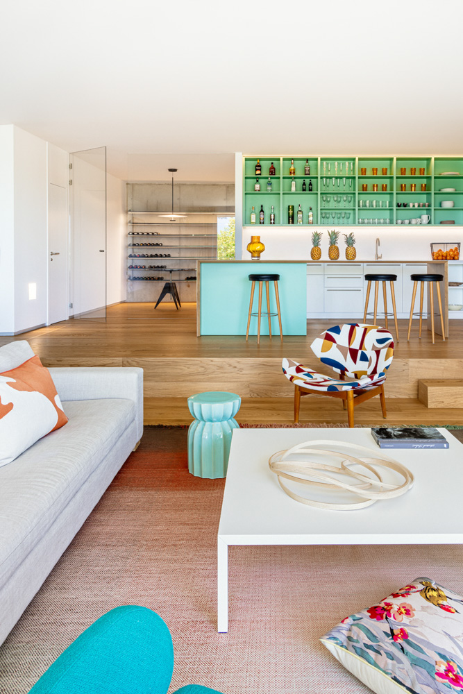 Salón moderno con sofá gris, alfombra bicolor, mesa blanca y cocina al fondo con estanterías verdes abiertas.